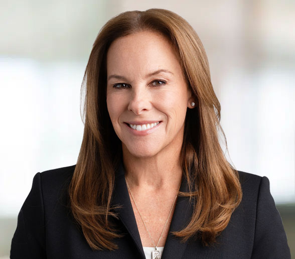 A woman with straight, shoulder-length brown hair, wearing a dark blazer and a necklace, smiles at the camera against a softly blurred background.
