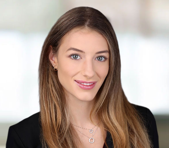A young woman with long, straight light brown hair, wearing a black blazer and necklaces, smiles at the camera against a blurred light background.